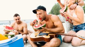 Muscular man eating a watermelon on the beach with his friends