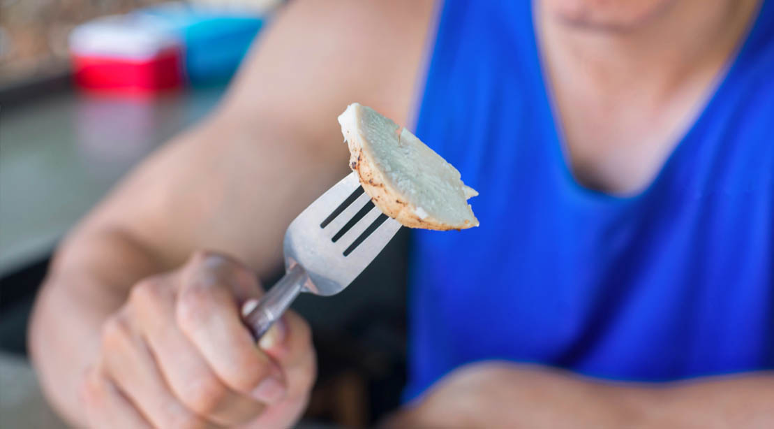 Fit man wearing a blue tank top eating grilled chicken with a fork