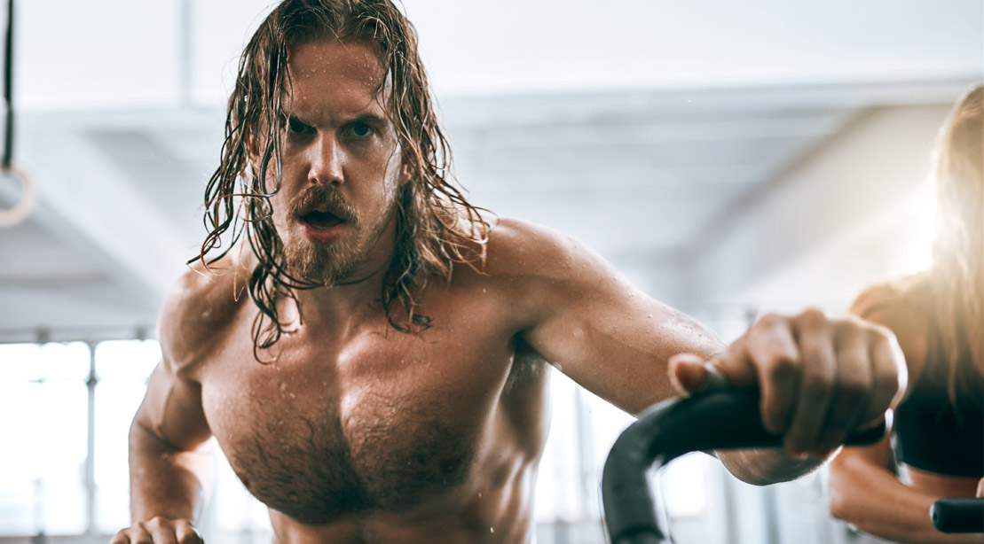 Sweaty muscular man with long hair doing cardio on an exercise bike