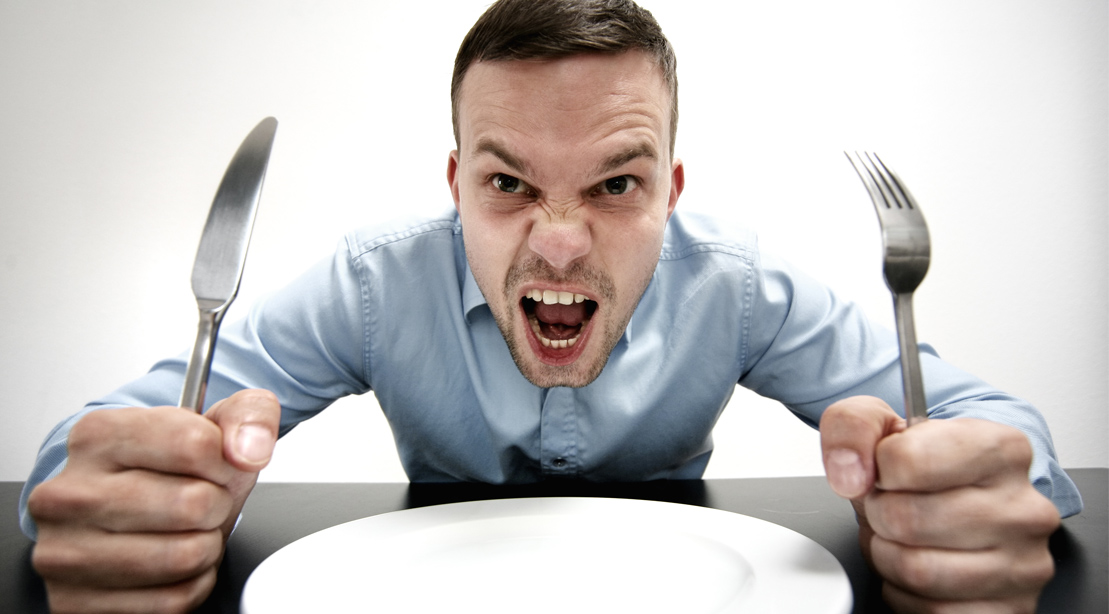 Angry man hungry and screaming while holding a fork and knife in front of an empty plate
