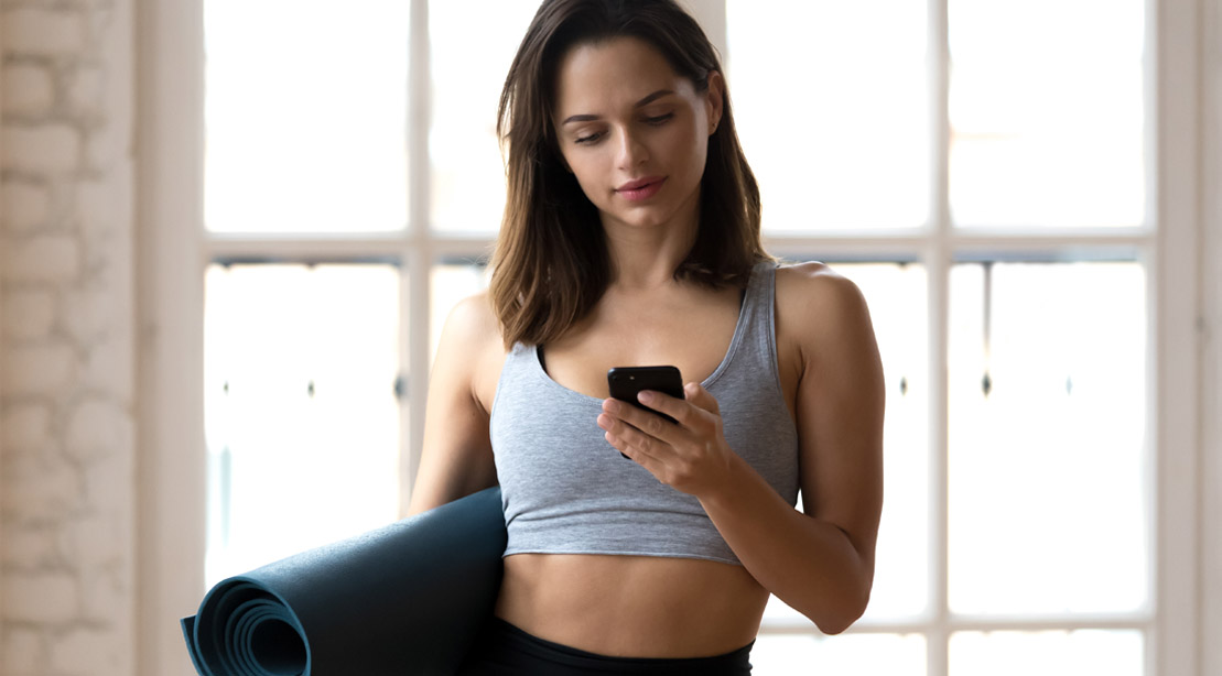 Female preparing for a home yoga workout using a health and fitness app while holding a yoga mat and smart phone