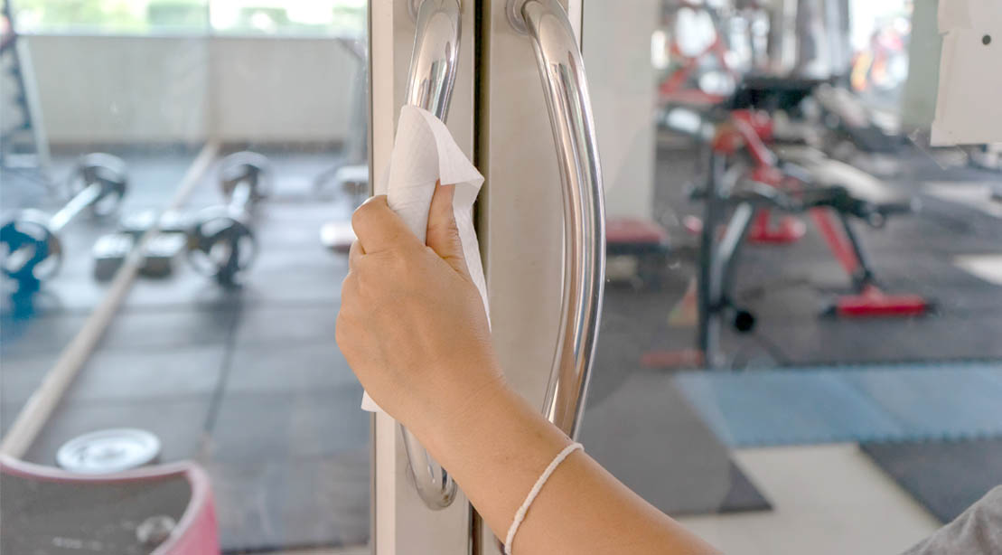 Female wiping down the handle of a glass door that leads to an empty gym