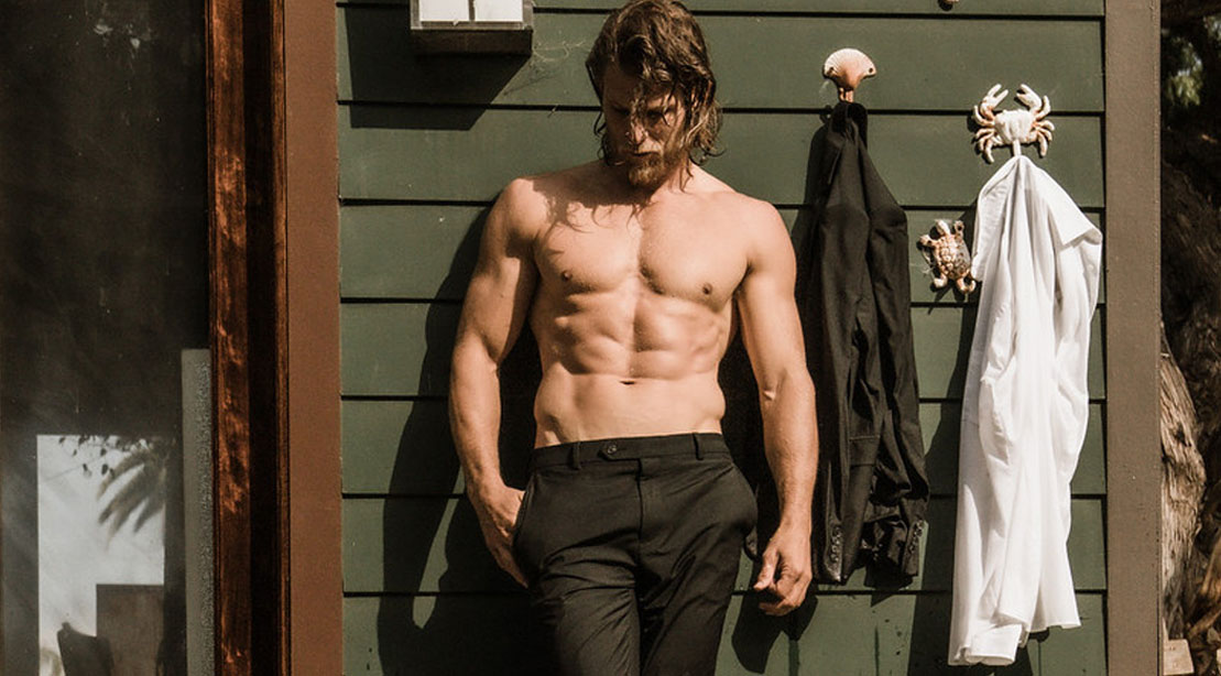 Travis Van Winkle posing against a house with his shirt off and ab muscles
